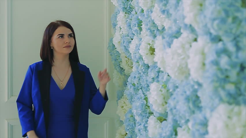 Beautiful stylish brunette woman with long hair in blue dress is walking next to flower wall at summer day. slow motion