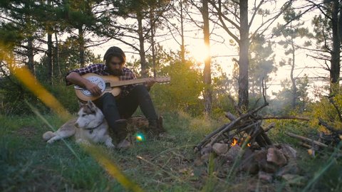 man playing guitar sitting with siberian husky dog near the campfire in forest during sunset 