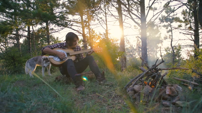 man playing guitar sitting with siberian husky dog near the campfire in forest during sunset 