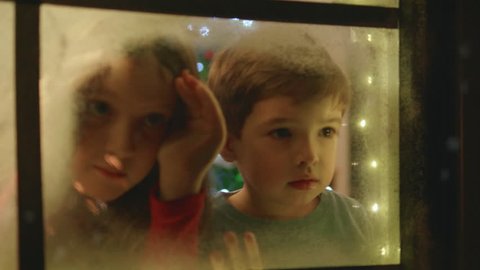 Adorable little boy and girl looking through the window and admiring first snow flakes and fireworks on Christmas evening.
