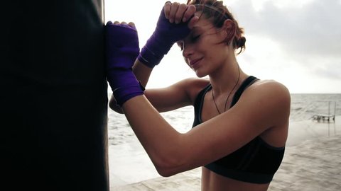 Closeup view of a young woman having a break after hard training by the boxing bag against the sun. Her hands are wrapped in purple boxing tapes. Training by the beach in summer