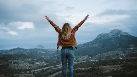Camera follows hipster millennial young woman running up to road side on top of mountain summit, jumps on top of rocks, raises arms into air, happy and drunk on life, youth and happiness