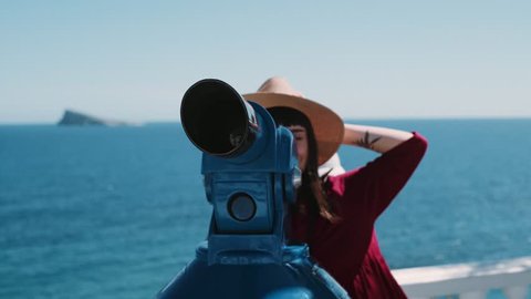Beautiful hipster millennial woman in red dress and straw hat looks through pay per view telescope on beach promenade, summer breeze blows her hair, she smiles and laughs enjoying holidays