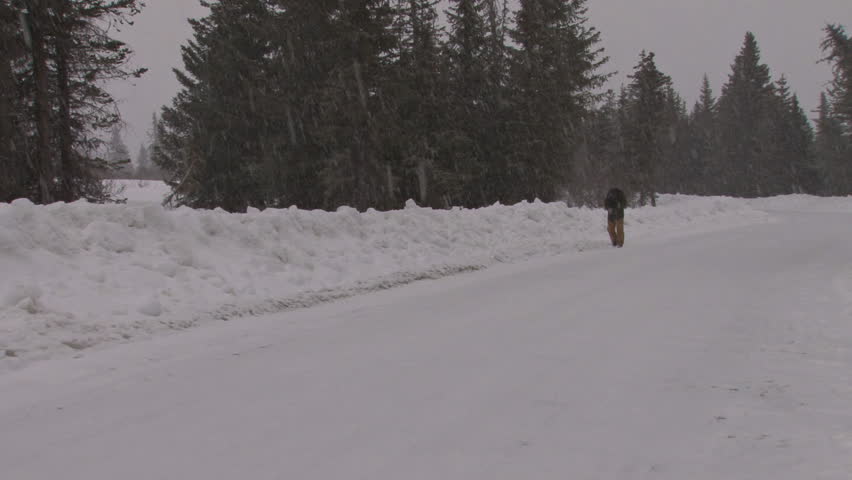 Cold, tired-looking man in winter gear stumbles along the side of a snowy road