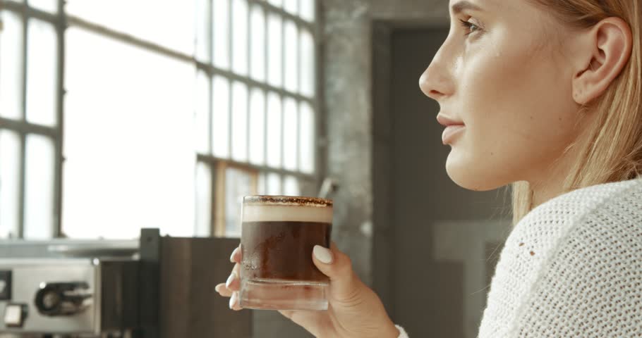 Attractive young woman wearing a white sweater in a loft style bakery or coffee shop with an ice latte in a glass