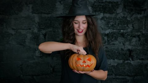 Halloween. Cute girl shows a cheerful witch. She is dressed in a black dress and hat. Girl holding pumpkin with a burning candle.