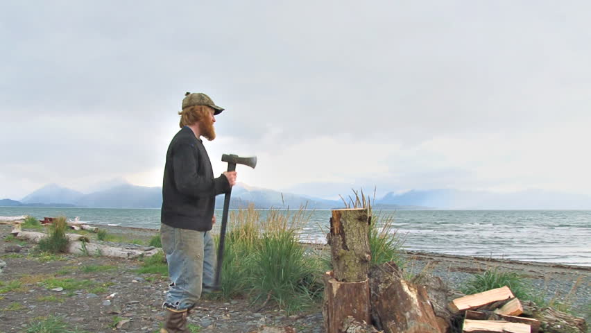 Rough and ready Alaskan man with a hat and beard splitting firewood at the beach