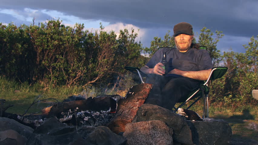 A man hanging out by a campfire with a beer responds to something and laughs.