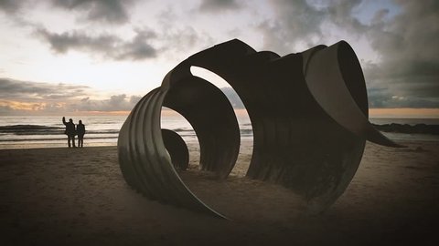 Young couple hold hands and kiss while watching sun set on a warm summer night in North West England. Giant sculptured steel shell on beach in Cleveleys, Blackpool