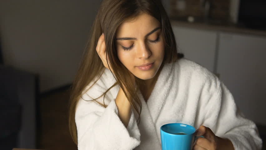 Beautiful caucasian girl, in a white bathrobe, enjoys morning coffee time in the kitchen, slow motion