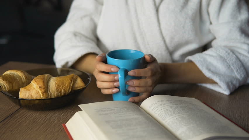 Young and beautiful caucasian girl, in a white bathrobe, dreaming while having coffee, morning time with croissants and a good book, slow motion