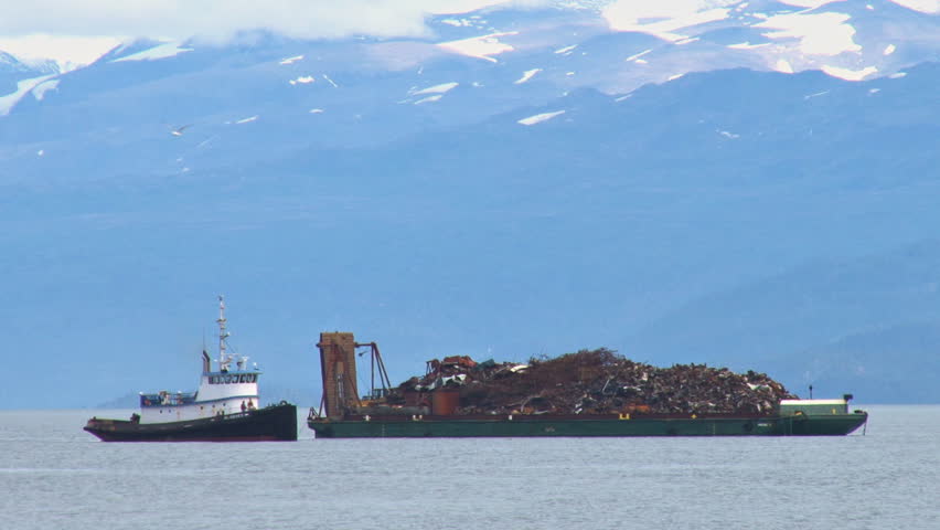 KACHEMAK, AK - CIRCA 2012: A tug boat moves past the barge piled high with scrap