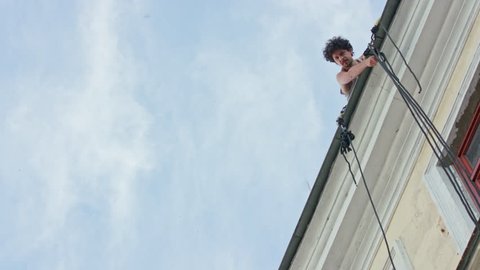 Warszaw, Poland - July 2017: Mattatoio Sospeso performes aerial dance show Out on the rope during Festival Sztukmistrzow. Sits at the edge of roof and waves hand. Sunny. Afternoon. Medium. Stabilized