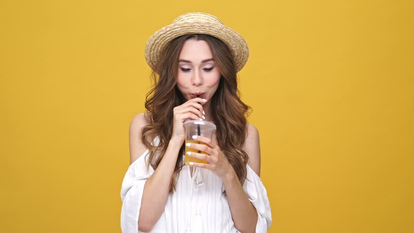 Pretty brunette woman in dress and hat drinking juice and dancing over orange background | Shutterstock HD Video #31321105