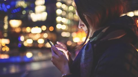 Woman Using SmartPhone at Night in Big City. 4K. Attractive young Woman texting, communicating on cellphone outdoors. Millennial Girl with phone, blurred Street skyscrapers and traffic lights.