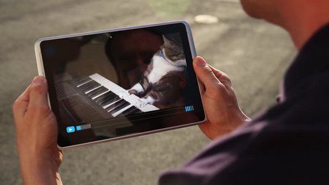 A man holding a tablet PC watches a viral video of a funny cat playing a keyboard or electric organ.	 	