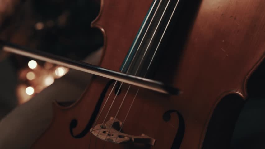 Young caucasian man with ponytail in plaid shirt playing cello with fiddlestick in room with christmas decorations