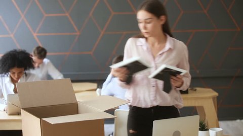 Fired young sad woman packing box standing near work desk, taking all her belongings after being dismissed resignation, upset employee quits from job collecting personal stuff at workplace to leave