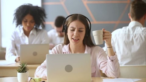 Funny happy young woman singing while listening to loud music in headphones sitting at office desk with laptop surrounded by diverse colleagues, lazy worker wasting time at workplace in co-working