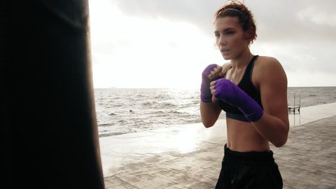Closeup view of a young woman training with the boxing bag against the sun. Her hands are wrapped in purple boxing tapes. Training by the beach in summer. Slowmotion
