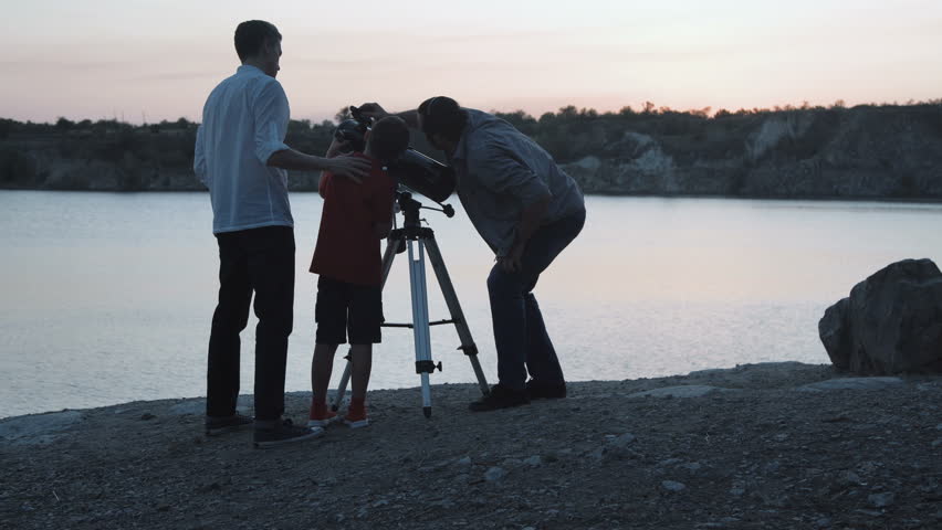 Two men and boy using telescope watching stars on shore of lake in twilight.
