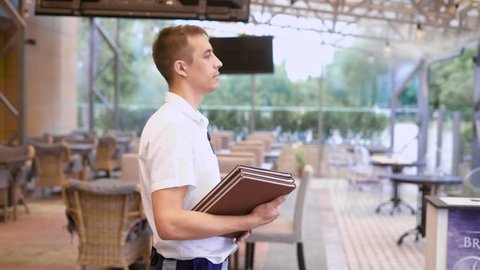 Close up of young male waiter standing in restaurant with menu and welcoming guests. Happy family coming in cafe for spending time together. Father, mother and son going and sitting down to the table.