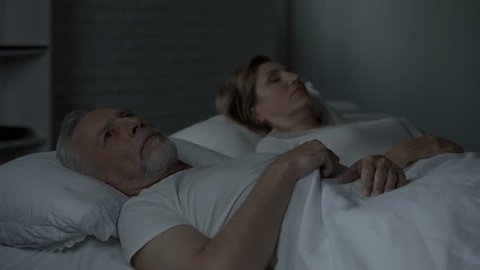 Elderly male in bed looking at female sleeping near, turning back to her, fight