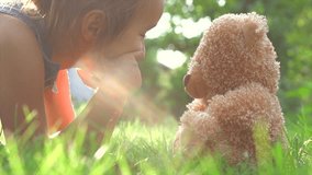 Little cute girl playing with her teddy bear in summer garden. Laughing child sitting on green grass with her toy. Happy childhood concept. Birthday gift. 4K UHD video 3840X2160 slow motion 240 fps