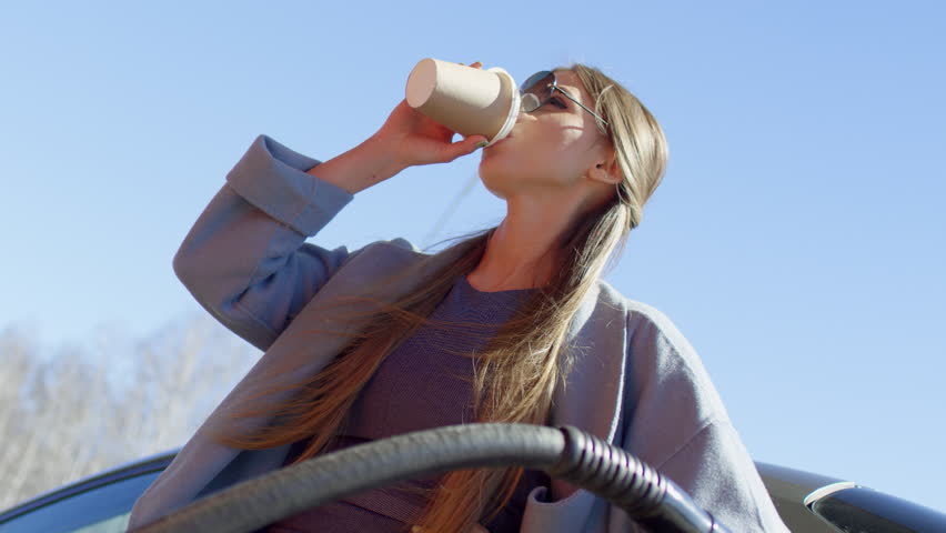 Low angle tilt down of beautiful young lady in sunglasses drinking coffee from disposal cup and smiling while filling up car at gas station at sunny day