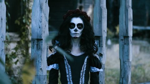 The Mexican Day of the Dead. The portrait of young woman with frightening skull make-up for Halloween on the background of wooden old house. 4K
