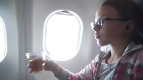 Young girl with glasses and headphones watches a video on the monitor built into the armchair and drinking juice in the cabin of the airplane stock footage video.