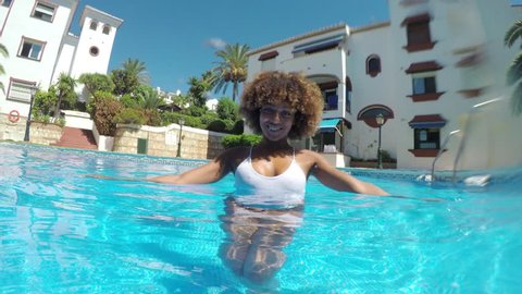 Attractive ethnic woman with curls wearing white swimsuit and posing in blue pool water looking at camera provocatively.