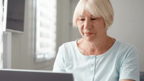 Senior woman sitting at home with laptop. Using computer talking via messenger. Smiling waving hand in greeting. Technology and communications use in active modern lifestyle of older people