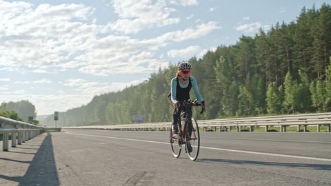 PAN of female cyclist in protective helmet and sunglasses riding bicycle along empty freeway surrounded by green forest