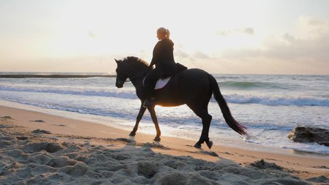 Young beautiful blonde woman in black clothing is carefully riding black horse on the seashore. Slow motion. Autumn sunrise or sunset on sea beach. Beautiful scene.