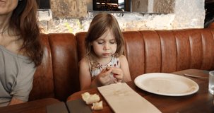 4k video. Four years age blonde girl eating olive with hands next to woman mother sitting in brown leather sofa at restaurant
