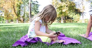 4k video. Four years age blonde girl watching smartphone, next to woman, sitting in towels on green grass in park of Retiro, in Madrid, Spain
