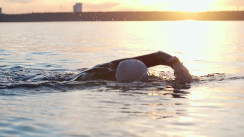 Tracking of woman in cap and goggles swimming in open water at sunset
