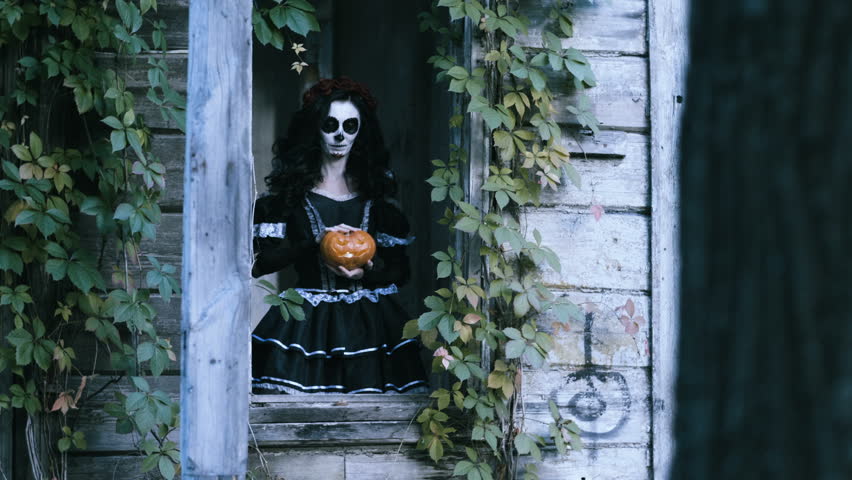 The Mexican Day of the Dead. The young woman with scary skeleton halloween make-up standing in the window of the old wooden house and holding a pumpkin with a lighted candle. 4K