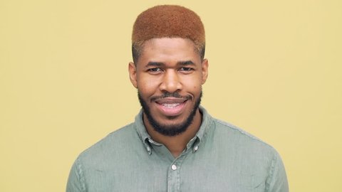 closeup dark-skinned guy with stylish haircut in blue skirt looking down then looking at camera,smiling broadly with white teeth over yellow background slow motion. Concept of emotions