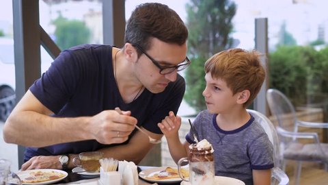 A young father tries his son's milk cocktail with ice cream and chocolate, the son laughs, the family is dining in the restaurant
