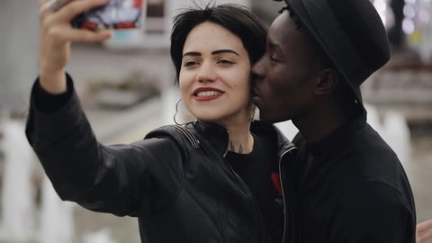 Slow motion of white woman and black man couple making a selfie video on smartphone. He kiss her cheek. They smiling, laughing together.