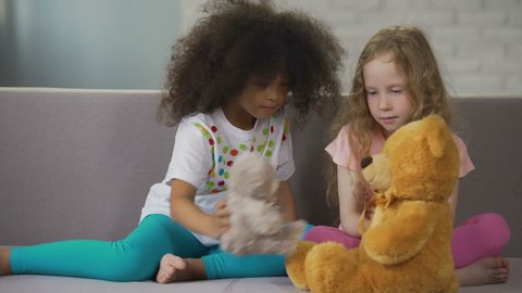 Two little multiracial girls sitting on couch and playing with teddy bears