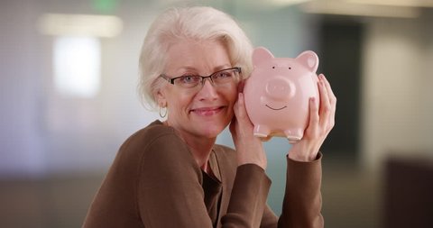 Cheerful senior woman shaking piggy bank smiling at camera. Portrait of mature woman saving money holding up piggy bank indoors. 4k