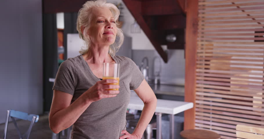 Slow motion shot of elderly woman enjoying glass of orange juice for breakfast inside kitchen. Cheerful mature woman drinking healthy glass of juice at home smiling. 4k