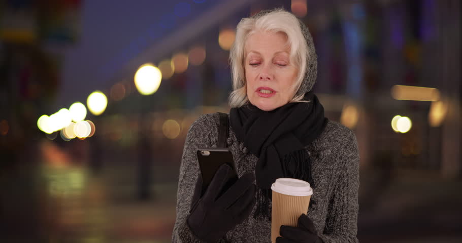 Senior woman removing glove with her teeth to text on smart phone in outdoor setting at night. Elder Caucasian woman in scarf and sweater using smart phone waiting for someone in evening. 4k