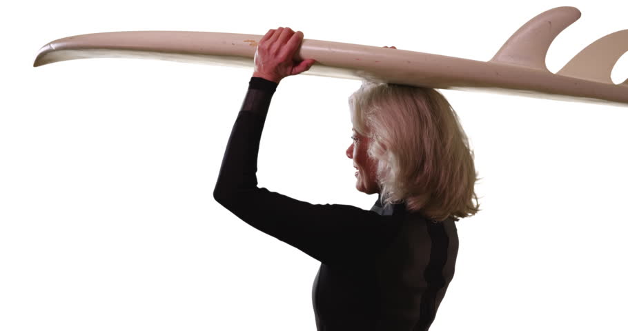 Attractive senior woman holding surfboard over head looking away from camera in studio. Elder woman on vacation posing with surfboard turning to smile at camera on white background. 4k