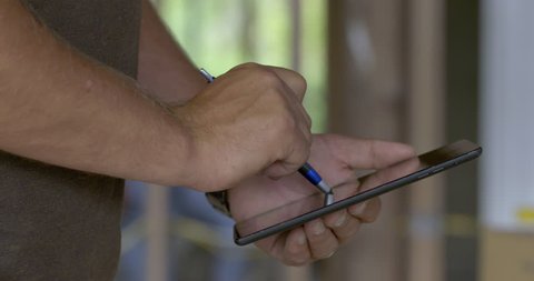 Detail of hands holding stylus and using tablet computer while contractor works on home under construction in soft focus background. Slider move 4K.