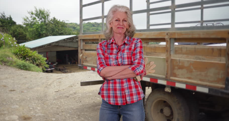 Senior woman in flannel shirt standing with arms crossed in outdoor country setting smiling. Portrait of elder woman posing by farm truck proud of family business. 4k