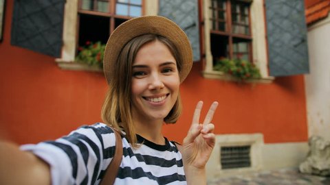Young attractive woman in a straw hat videochatting with friends, waving on the medieval street background. Talking happily about vacation trip on a sunny day.